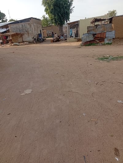The image depicts a sandy area in front of a row of buildings, possibly in Gulak, Nigeria. The buildings appear to be constructed from a mix of concrete, corrugated metal, and mud. One building has "DUHU" written on its facade. Several people are visible. In the left portion of the image, near a structure with a thatched roof, a person in red clothing is partially visible. Further to the right, three people are gathered near some logs, and another person in blue is walking past a fenced entrance. Two motorcycles are parked in front of one of the buildings. The ground is covered in loose sand and debris, with scattered vegetation in some areas. The sky is overcast, suggesting it might be late afternoon or early morning.