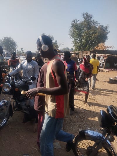 This image captures a daylight scene in Unguwan Rimi, Nigeria, depicting an unpaved, bustling outdoor area, likely a market or thoroughfare. The foreground is dominated by a young Black male, mid-stride towards the right of the frame. He wears white over-ear headphones, a sleeveless shirt (tan top, red bottom), and blue jeans, holding a yellow, rectangular object horizontally in his right hand.
To his immediate left, another Black male in a light-colored traditional tunic stands near a dark-colored motorcycle with chrome accents, its headlight and handlebars visible. Other individuals are dispersed throughout the mid-ground: a man in an orange and black striped shirt, a man in a dark blue short-sleeved shirt with a chest emblem, a woman in a red and white patterned head covering and matching dress looking towards the camera, and a man in a yellow shirt moving away from the viewer.
The ground consists of dry, reddish-brown dirt. In the background, rudimentary structures with corrugated metal roofs and brick or mud walls suggest market stalls or residential units. Several green trees with dense foliage are visible against a clear, bright blue sky, indicating a sunny day. Multiple motorcycles are present, either parked or in motion, contributing to the scene's dynamic atmosphere.
