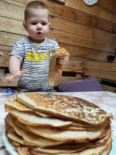 A young child, likely a toddler, stands at a table laden with a tall stack of golden-brown pancakes. The child is wearing a striped t-shirt with "HEY!" written in yellow letters across the chest. They are holding one pancake in their right hand, seemingly about to eat it or offer it. The child's expression is one of intense focus and perhaps a bit of surprise, with their mouth slightly open.
The setting appears to be a rustic interior, with wooden walls and paneling. A purple pillow rests on a bench behind the child. A clock is visible on the wooden wall above the child, its hands indicating a time between 5 and 6. The table is covered with a patterned tablecloth. In the foreground, the stack of pancakes dominates the lower part of the frame, showing their layered texture. The lighting suggests it might be evening or indoors. The watermarked text "HONOR 400" is visible in the bottom left corner. The scene evokes a sense of home and a simple, everyday moment centered around food and a curious child. The location context provided is Эрхирик, Russia.
