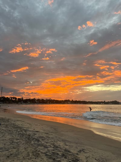 This is a vertical, full shot of a beach at sunset. The sky is a dramatic mix of gray clouds with vibrant orange and yellow streaks, indicating sunset or sunrise. The sun is low on the horizon, casting a warm glow over the scene. The ocean is calm with small waves breaking on the shore. A sandy beach stretches along the coastline, with footprints visible in the wet sand.
In the water, a single person is captured mid-air, appearing to be jumping or diving. This figure is silhouetted against the bright, reflective water, adding a dynamic element to the tranquil setting. Along the coastline, a line of palm trees and buildings suggests a populated area, possibly a town or resort. The overall mood is serene and awe-inspiring, characteristic of a beautiful sunset by the sea. The time of day is clearly sunset due to the colors in the sky and the low angle of the sun. The location is identified as Arugam Bay, Sri Lanka, a popular surfing destination. There is no visible text in the image.
