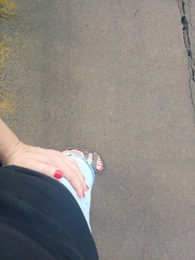 The image shows a person's legs and feet standing on a cracked asphalt surface. The person is wearing blue jeans and brown sandals. Their toenails are painted red. A black garment is visible in the lower left of the image, likely part of the person's clothing. The asphalt has some yellow discoloration on the left side and a prominent crack running vertically on the right. The scene appears to be outdoors, possibly in a street or walkway. There is no direct indication of the specific location within Trypillia, Ukraine.
