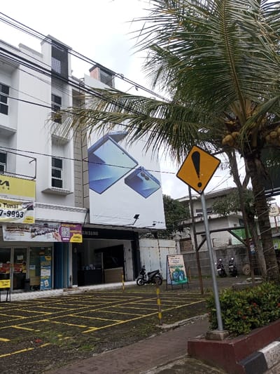 This image depicts an urban commercial scene in Kodarapalli, India, under an overcast sky. On the left, a multi-story white building features multiple dark-framed windows. A large banner showcasing two blue smartphone-like electronic devices is prominently displayed on the building's facade, directly above a storefront identified by a "SAMSUNG" sign. Below this, ground-level retail spaces are visible, one featuring an "AORA TUNAI" sign with a phone number and another displaying a QR code and promotional material. In the mid-right, a yellow diamond-shaped warning sign with a black exclamation mark is mounted on a gray pole, positioned next to a tall palm tree laden with green coconuts. Two motorcycles are parked to the right of the building, one black and one light-colored, near an A-frame sign with green graphics. The foreground consists of a paved, partially overgrown area marked with yellow grid lines, suggesting parking spaces. A small planter containing green bushes with red flowers is visible in the lower right. Overhead, numerous electrical wires traverse the scene. No individuals are present, and no actions or interactions are depicted.