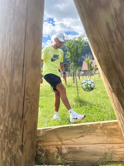 A young man in athletic wear, including a yellow Nike t-shirt, black shorts, white socks, and white sneakers, practices juggling a soccer ball in a grassy outdoor area. The man is positioned to the left of the frame, looking down at the ball, which is in mid-air. He is framed by two large wooden posts. In the background, there are trees, houses with red roofs, a fence, and mountains under a cloudy sky. The scene appears to be in Gusinje, Montenegro, a mountainous region.