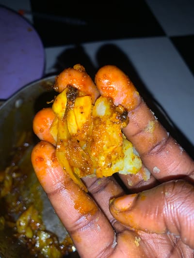 A close-up shot shows a person's hands holding a piece of food. The food appears to be a mixture of potato and possibly some type of meat or fish, coated in a reddish-orange spice blend. The hands are dark-skinned and appear to have spices on them as well. The background is blurred, with a hint of a black and white checkered pattern on the left and a purple object on the bottom left. The lighting suggests it might be indoors or at night. The overall impression is that someone is about to eat or is in the process of handling food, likely a meal prepared with spices. The texture of the food and hands is detailed, with visible grains of spice and skin texture. There is no visible text in the image.