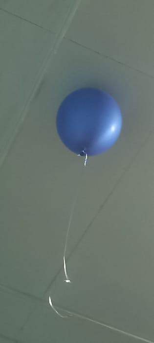 A single, inflated, spherical blue balloon is observed floating near a light-colored ceiling. The balloon has a reflective sheen on its upper surface, indicating an unseen light source. A thin, light-colored ribbon or string, appearing white or silver, is tied to the balloon's knot and hangs downwards, curving gently before exiting the bottom-left of the frame. The ceiling surface is light grey or off-white and features linear indentations or seams that suggest a panelized construction, with lines intersecting to form distinct sections. No individuals or additional objects are present in the scene. The image depicts an indoor environment located in Kyzyltepa, Uzbekistan.