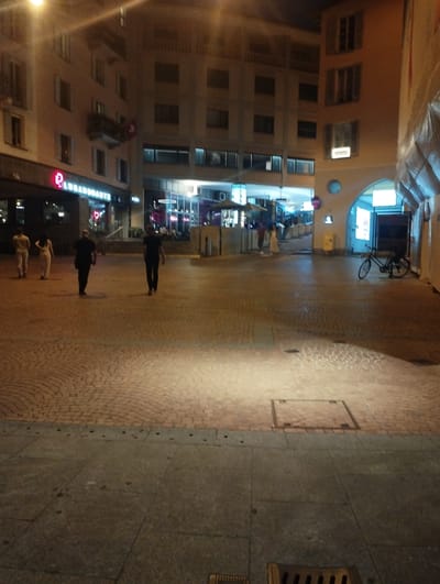 The image depicts a nighttime street scene in Lugano, Switzerland, likely in the city center. In the foreground, a cobblestone pavement transitions to a paved area with visible manhole covers and a dark grating. Two individuals are walking away from the viewer on the paved area, one wearing dark clothing and the other in lighter colored attire. Further in the midground, on the cobblestone section, two more figures are seen walking in the same direction, also away from the camera, dressed in light and dark garments respectively.
Behind the pedestrians, a building with illuminated signage that reads "P. LUGANO ARTE" is visible. The building itself has multiple floors with windows and balconies. Across the street, an arched structure leads into a dimly lit area with more signage and what appears to be a bicycle parked on the right. The overall atmosphere is nocturnal, with artificial lighting casting shadows and illuminating parts of the urban environment.