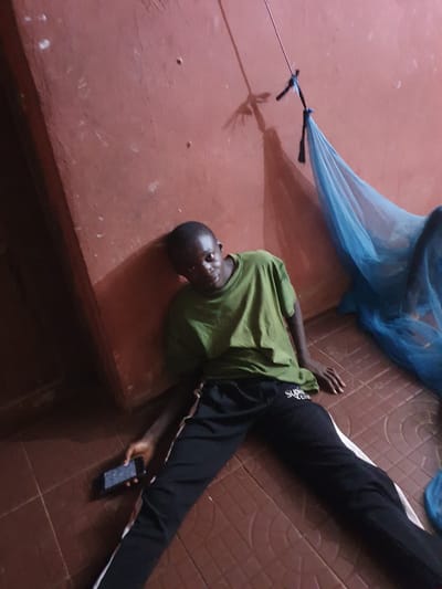 A young person, likely male, with dark skin and short dark hair, lies on their back on a reddish-brown tiled floor, partially resting on a lower reddish-brown platform or bench. They are wearing an olive green short-sleeved t-shirt and black track pants with a white vertical stripe down the side. A dark-colored smartphone is held in their right hand. Above and to the right, a light blue, sheer fabric, resembling a mosquito net, is draped. A thin metal rod or wire is visible, connected to the blue fabric. The overall lighting is dim, indicating an indoor setting, possibly in the evening or a poorly lit room in Idah, Nigeria. The person looks directly at the camera with a neutral expression. Visible text on the left leg of the track pants appears to read "SKATE CRU".