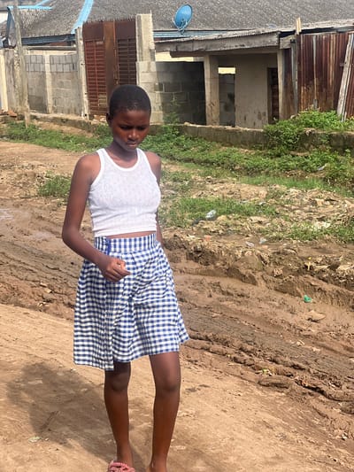 A young person stands outdoors in a bright, sunlit environment, likely in Ijegun, Nigeria. The individual has dark skin and short, dark hair, and is wearing a white tank top with a subtle textured pattern and a blue-and-white checkered skirt. Their eyes appear closed or heavily downcast, and one foot with a pink sandal is visible.

The setting is characterized by a dirt ground, which is uneven and appears muddy in places, showing tire tracks and scattered debris or rubbish. Green vegetation, consisting of weeds and grass, grows alongside the muddy path.

In the background, there are several simple buildings constructed from concrete blocks, some appearing unfinished or in disrepair. These structures feature corrugated metal roofs, with one roof having a small blue satellite dish attached. The overall scene suggests a rural or semi-urban area with basic infrastructure, typical of parts of Nigeria. The time of day is clearly daylight, indicated by the strong illumination and visible shadows. There is no visible text in the image.