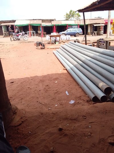 The image depicts an outdoor commercial area in the city of Ankpa, Nigeria, under bright daylight. The foreground consists of reddish-brown dirt ground. On the far left, a portion of a dark-clad leg and a dark sandal are visible, suggesting the photographer's low perspective. A large stack of approximately twenty to twenty-five grey, cylindrical pipes lies horizontally across the right foreground on the dirt. Scattered pieces of white plastic litter are visible on the ground near the pipes.
A dirt road separates the foreground from the background structures. On the left side of this road, two men are standing near a red and chrome motorcycle. One man, wearing a dark top and light-colored trousers, stands facing a second man, who is dressed in a dark top and camouflage-patterned trousers. In the center of the dirt road, a small brick structure with a dark pole is visible. Further to the right along the road, a dark blue station wagon is parked, facing left, partially obscured by a wooden table holding various items and several white sacks stacked behind it.
The background features a continuous row of single-story buildings with corrugated metal roofs, functioning as shops or stalls. Many of these buildings are decorated with green and red fabric drapings. A signboard with white text on a dark background is visible on one of the buildings. Sparse green trees rise above the rooftops against a bright, hazy sky. A red and yellow architectural element is present on the far right edge of the frame.