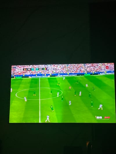 The image displays a television screen showing a soccer match between Belgium (BEL) and Senegal (SEN). The score is 0-0, and the game is at the 17:44 mark. The screen is framed by a dark border, suggesting it is a television within a room. The soccer field is bright green with white lines marking the boundaries and center circle. Several players in white and green uniforms are visible on the field. In the background, a stadium filled with spectators wearing various colored clothing can be seen. Advertising banners are present along the sideline. The scene is captured in Gwarinpa, Nigeria, indicating the television is located in that city.