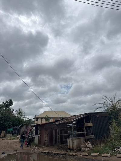 The image depicts a street scene in Ihiagwa, Nigeria, under a heavily overcast sky. A dilapidated corrugated metal structure stands to the right of the frame, with evidence of makeshift construction and various objects stored within. To the left, a taller building can be seen partially obscured by the metal structure. At least three people can be seen walking along the unpaved road. The road appears to be wet, possibly due to recent rain. There is vegetation and a palm tree visible. Electrical wires stretch across the sky. The environment suggests a low-income or underdeveloped area.