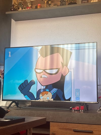 A television screen, oriented vertically within the frame, displays an animated cartoon scene. The prominent character on screen is a large, muscular figure with dark hair, a black mask covering the upper face, and a determined expression, wearing a dark blue and light blue suit. Beside this figure is a smaller, child-like character with light brown hair and wide blue eyes, also in a dark blue outfit, looking surprised or curious. Faint vertical light reflections or stripes are visible across the screen. In the bottom left corner of the screen, the number "1" is displayed, and in the upper right, small icons resembling a safe, a grid, and a circle are present.
The TV is situated indoors, likely in a living area or kitchen in Kuklen, Bulgaria. To the left of the screen, a textured, light gray wall is visible, alongside a dark gray shelving unit holding various decorative items, including bottles, small figurines, and dried plants. A "VERSACE" label is partially seen on an item on the shelf. To the right of the TV, a wooden cabinet or counter with a long, vertical metal handle is visible. Several small red electrical components or devices are attached to the right side of the TV's frame, with cables running behind it. The indoor setting is brightly lit, suggesting daytime.