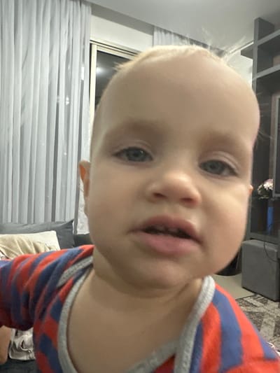 A close-up, waist-up shot shows a young child with blonde hair and blue eyes. The child is wearing a red and blue striped shirt with a grey trim. The background features sheer white curtains, a window, and part of a living room with a couch, pillows, and a shelving unit. The child's face is turned towards the camera, with their mouth slightly open. The lighting appears to be indoor, possibly from a flash. The scene is consistent with a residential setting in Florianópolis, Brazil.