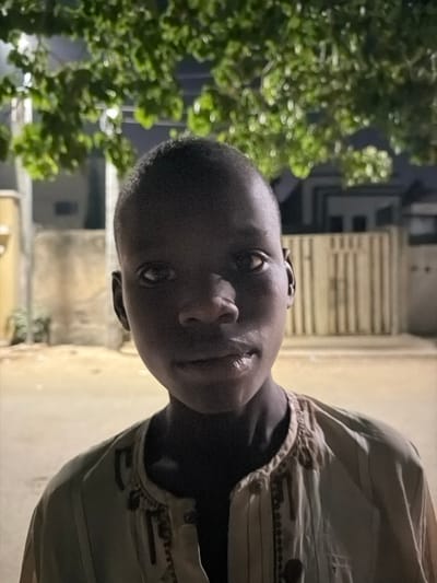 The image presents a close-up depiction of a young male, estimated to be between 10 and 14 years of age, situated outdoors at night in Gandu Albasa, Nigeria. The individual, possessing dark skin and short, dark hair, is positioned centrally in the frame, directly facing the camera with a neutral expression. He is wearing a light-colored, possibly beige or light brown, long-sleeved garment featuring distinct dark embroidered patterns along its front placket. Direct artificial illumination highlights the subject's face and upper torso. The background is out of focus, revealing green foliage from an overhead tree at the top of the frame. Beyond the subject, indistinct residential structures are visible, including a light-toned building on the left and a darker structure with vertical elements, consistent with a fence or gate, on the right. A dimly lit ground surface is visible below these background elements. No other individuals, explicit actions, or direct interactions beyond the subject's gaze at the camera are observed.
