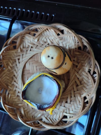 A top-down shot captures two small, decorative items resting within a woven bamboo basket. The basket has a circular base with an intricate crisscross weave. The larger object, positioned towards the top right, is an oval-shaped, light-colored object with dark brown markings that resemble a face. It has two small dark holes, possibly representing eyes, and appears to be made of ceramic or a similar material. To the left and slightly below this, is another object that looks like a small, open bowl or container. This container has a dark brown rim with a yellow cord or trim encircling its opening. Inside, it is filled with a fluffy, white material. The basket itself is placed on a dark, reflective surface, which could be a stovetop, given the textured black element visible at the top of the image, suggestive of oven vents. The overall scene is illuminated by overhead lighting, casting subtle shadows. There are no people or animals present, and no discernible interaction between the objects. The location is not specified within the city of Mykolaiv, Ukraine, and is thus not described.