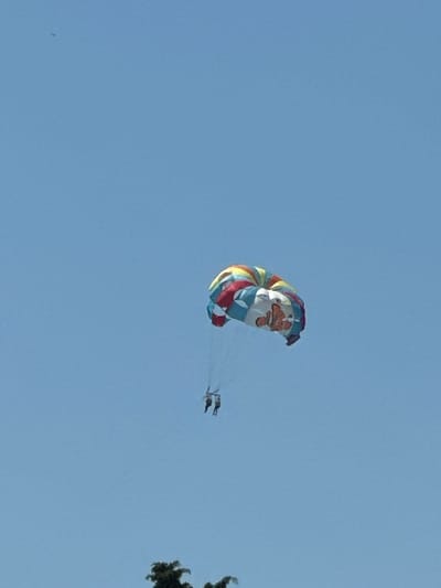 The media file shows a bright, clear sky over Boreti, Montenegro, with a parasail in the air. The parasail is colorful, with stripes of red, yellow, blue, and white, and features a depiction of Santa Claus on its fabric. Attached to the parasail are two people, appearing to be enjoying a recreational activity. One person is directly beneath the parasail, while the second person is slightly to the right, trailing behind. A portion of a green tree is visible in the upper right corner of the frame, suggesting an outdoor location, possibly near a coast or resort area. The lighting indicates it is daytime, likely during good weather, given the clear blue sky. No text is visible in the media file.