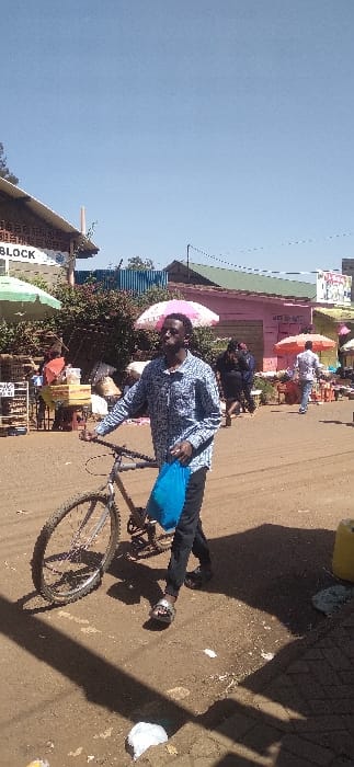 A man rides a bicycle on a dirt road. He wears a patterned shirt and dark pants, carrying a blue bag. Several people are visible in the background under colorful umbrellas, likely at a market. A building with signage is on the left. The sky is clear. The overall impression is of a busy street scene in a less developed area.