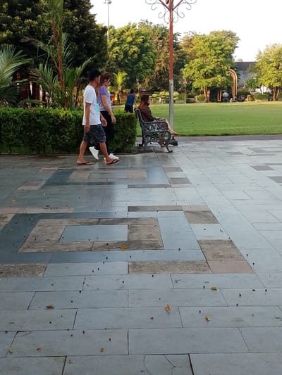 Three people are walking on a tiled pathway in the city of Pati, Indonesia, located at the coordinates -6.753, 111.04. Two of the people are walking side by side, and the third is sitting on a metal bench to the right. The person on the bench is wearing a purple shirt. The people on the pathway are both wearing light-colored clothing. The scene takes place in an outdoor park with green grass and trees.  There are palm trees and bushes bordering the path. There is a lamp post in the center of the image, and another lamp post is visible in the background.  The bench is empty except for the seated person. The pathway is surrounded by a tiled area. The path is made of large, rectangular tiles with a pattern of smaller, square tiles.  The sky is clear and blue, and the sun is shining.  There is one person in a blue shirt walking in the background to the right.  The scene appears to be taking place in a public park.  There is some debris on the ground.