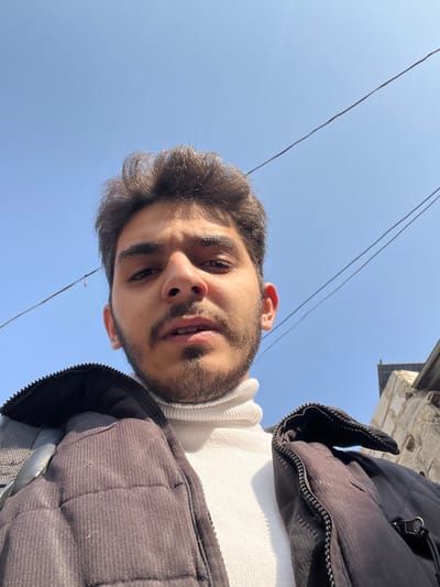 The image presents a low-angle, head-and-shoulders portrait of a young adult male against a clear blue sky. The individual has dark hair, a short beard, and a mustache. He is wearing a white ribbed turtleneck sweater and a dark, possibly black, quilted jacket. A grey backpack strap is visible over his left shoulder. His mouth is slightly open, and he is looking directly into the camera. Two dark utility wires traverse the upper portion of the frame against the sky. A partial view of a light-colored building structure with a darker roofline is visible on the far right edge. The scene is illuminated by bright daylight and is located in Rukn ad-Din Municipality, Syria.