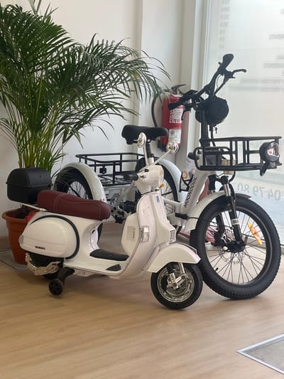 The image shows a white scooter and a black electric bicycle parked indoors, likely in a retail setting or showroom. The scooter, which resembles a Vespa, has a prominent headlight, chrome handlebars, and a brown seat. It is positioned in the foreground, with a black storage box attached to the rear. The electric bicycle is behind the scooter and is black with a large, knobby tire. It features a front rack with a basket and a headlight. A red fire extinguisher is visible near the handlebars of the bicycle. A lush green plant in a brown pot is in the lower left corner of the frame. The background consists of a white wall with some blurry text, possibly indicating a store or office. The lighting suggests it is daytime, and the setting is indoors, as indicated by the flooring and the absence of natural light cues like shadows from windows. The overall impression is one of a display of electric vehicles. The word "MOONCOOL" is visible on the frame of the electric bicycle.