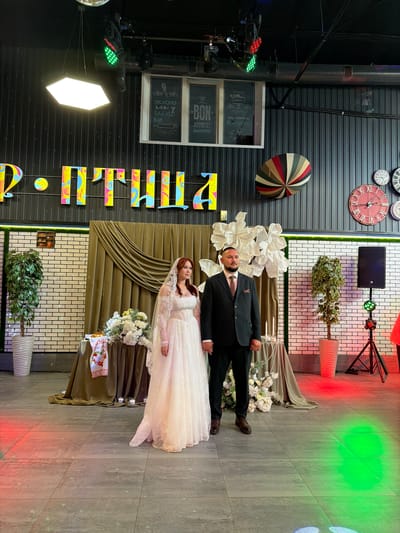 A wedding photo is taken of a bride and groom standing side-by-side in front of a draped background. The bride is wearing a white lace wedding dress and a veil, while the groom is in a dark suit and tie. They are holding hands. The room appears to be a banquet hall with decorative elements such as illuminated letters on the wall, large paper flowers, clocks, and a decorative umbrella. There are also potted plants, a speaker on a stand, and colored stage lights casting green and red hues on the floor. The floor is tiled. The lettering on the wall is in Russian and likely spells out "Свадьба" (Wedding) or a similar celebratory phrase. The presence of multiple clocks on the wall, particularly the large red-faced one, could be a reference to time or a nod to the city's history if it's a local landmark. The overall impression is a festive and decorated wedding reception venue, possibly in Chernushka, Russia, given the Cyrillic lettering.