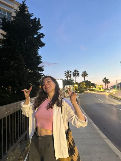 A person is standing near a street in Bečići, Montenegro. The location is identifiable by the presence of palm trees and a building with a European architectural style with barred windows. The person is giving a peace sign to the camera, visible at the bottom left of the frame. They are wearing a pink crop top and white jacket. A leopard print bag hangs around their shoulder. Graffiti is seen on the metal bars where the person stands. A street with sparse traffic is seen in the background. The lighting indicates it may be evening. The sky shows a gradient of blue transitioning to dusk colors.