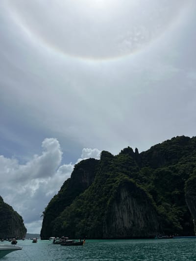 An atmospheric halo, likely a sun halo, is visible in the sky above a seascape featuring lush green karst formations. The halo is a circular or arc-shaped optical phenomenon, appearing as a bright ring around the sun. The water is a clear turquoise, dotted with numerous longtail boats, common in Ao Nang, Thailand. Some of these boats appear to be carrying passengers, and at least one boat has people standing on it. The foreground shows a portion of a white boat. The background is dominated by steep, tree-covered cliffs. The sky is partly cloudy, with patches of blue sky peeking through. The overall impression is a serene, tropical scene with a natural atmospheric spectacle.