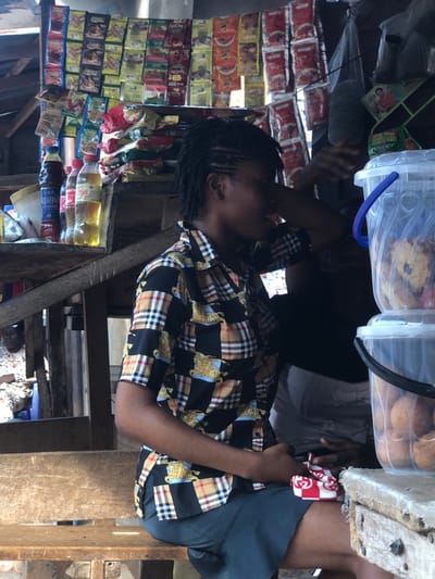 A woman sits, turned to the left, near a stand filled with small goods in red and yellow packaging. She wears a checkered shirt with images of horses and is holding something in her hands. Above the stand are transparent plastic containers filled with baked goods. Someone stands to the right of the woman wearing white shorts, and to her left, someone appears to be holding a microphone. The setup appears to be outdoors.