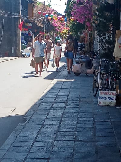 The image depicts a street scene in Hoi An Ward, Vietnam, likely a tourist area, characterized by a narrow street lined with buildings and adorned with vibrant flowers. A group of people are walking along the cobblestone sidewalk. In the foreground, a man wearing sunglasses, a light blue short-sleeved shirt, and khaki shorts is walking with a brown shopping bag in his left hand. To his right, another man in a light brown short-sleeved shirt and white shorts is walking. Behind them, a woman in a white t-shirt and a patterned skirt is walking, carrying a similar brown shopping bag. Further back, several other individuals are visible, some appearing to be tourists.

On the left side of the street, shops are visible with merchandise displayed. One shop has a red flag hanging. The street is decorated with colorful lanterns strung overhead. In the background, dense pink bougainvillea flowers cascade down from above, adding to the picturesque atmosphere.

To the right of the sidewalk, several parked motorbikes and bicycles are present. One of the bicycles has a sign attached to its basket that reads "CHO THUÊ XE ĐẠP BICYCLE - FOR RENT" followed by "LH: 0971403528". The architecture suggests a historic district, with textured buildings and a paved walkway. The lighting indicates it is daytime with bright sunlight casting shadows on the ground.