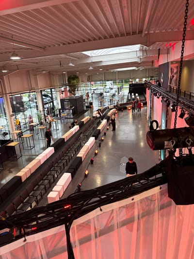 The image displays a large, high-ceilinged indoor event space in Herentals, Belgium, configured for a corporate or product launch event. The venue features an industrial-modern aesthetic with exposed metal ceiling trusses, ductwork, and integrated overhead lighting.

A central aisle-like pathway is established, flanked by curved rows of seating composed of black chairs interspersed with alternating black and light beige rectangular poufs or benches. This aisle is bordered by a series of floor-level amber/red spotlights.

On the right side of the frame, a stage or presentation area is visible, partially obscured by foreground elements. It incorporates white translucent drapery illuminated by red uplighting and overhead professional stage lighting fixtures mounted on a truss system. A large backdrop on the stage and a projection on the polished concrete floor prominently display a Mercedes-Benz star logo.

Approximately 15-20 individuals are present throughout the space. Most are dressed casually, with some wearing dark professional attire, suggestive of event staff. Actions observed include individuals standing, conversing in small groups, walking, and one person seated. A person in a black top and light trousers stands in the foreground, facing away from the camera. Another person in a black shirt is positioned near a grouping of high-top tables.

To the left, near a wall of large, floor-to-ceiling windows, several high-top cocktail tables with light wooden tops are arranged. A dark, modular counter unit with "TrueVision" branding, potentially a bar or registration desk, is also situated in this area. A tall potted tree serves as decor. The windows offer a view of an outdoor paved area with parked vehicles.

The immediate foreground features a structural element with a dense bundle of black cables, a suspended black lighting fixture, and white translucent fabric back-lit with red illumination. The polished concrete floor is visible throughout the main area.