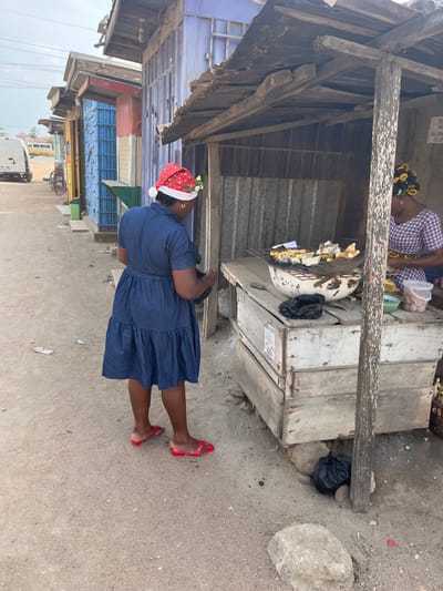 The image depicts a daytime street scene in Ngleshi Amanfro, Ghana. In the foreground, a woman stands with her back partially to the viewer, facing a street vendor stall on the right. She wears a blue denim short-sleeved dress, a red Santa hat with white trim and green festive decorations, and red slide sandals. Her left arm is slightly bent, and she appears to be interacting with the stall.
To her right, a makeshift vendor stall is constructed from raw wooden planks and corrugated metal sheeting, topped with a weathered corrugated metal roof supported by wooden posts. Within this stall, a second woman is visible on the right side, wearing a patterned top and having yellow hair rollers in her dark hair. She is attending to food items on a wooden counter.
On the stall's counter, a metal grill holds several pieces of food, likely plantains or yams, positioned above a metal basin. Nearby, clear plastic containers hold small, light-brown food items, possibly groundnuts, and a black plastic bag rests on the counter. Another black plastic bag is on the ground near the stall's base, alongside a large rock. The ground consists of light-colored dirt and gravel, with scattered small pieces of refuse.
In the background, a row of similar, brightly colored corrugated metal structures, likely additional stalls or shops, lines the unpaved street. A white van is visible parked further down the road on the left. The sky is overcast.