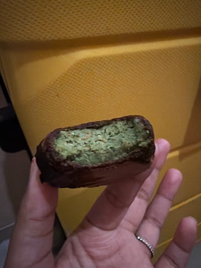 A close-up shot shows a person's hand holding a cut-open, chocolate-coated food item with a green filling. The background is a textured, bright yellow surface, possibly a piece of luggage or furniture. The food item appears to be a cookie or a candy bar with a cake-like, green interior. The person's hand is visible, with a ring on one finger. The image was taken in East Jakarta, Indonesia.