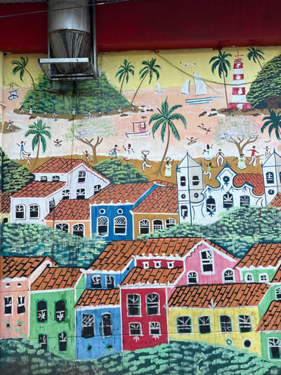 The image shows a vibrant mural painted on a wall. The mural depicts a coastal town scene with colorful houses, palm trees, sailboats, and a lighthouse. In the background, there are hills and a clear sky. People are depicted engaged in various activities, such as carrying baskets and dancing. A large metal pipe hangs from above, partially obscuring the top of the mural. The mural is located on the wall of a building in São Paulo, Brazil, likely in a neighborhood known for its street art.