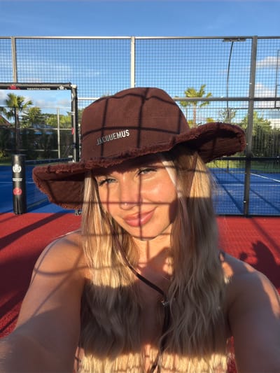 The image depicts a young woman wearing a brown bucket hat with the word "JACQUEMUS" embroidered on it, and a fuzzy brim. She is in an outdoor setting that appears to be a sports court, possibly for padel tennis, with a blue surface and red outer areas. The court is enclosed by a wire fence, and palm trees are visible in the background, suggesting a tropical or subtropical location. The sky is clear and blue, with the sun casting strong shadows across the scene, including on the woman's face. The woman has blonde hair and is looking towards the camera with a smile. There is a black cylindrical object with logos on it near the fence on the left side of the frame. The overall lighting suggests it is late afternoon or early morning. The location is identified as Thep Krasatti, Thailand.