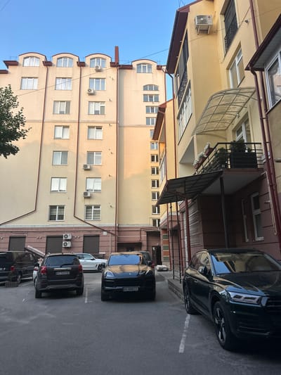 The image depicts a residential building in Ternopil, Ukraine, with a parking lot in front. The building is light beige with maroon accents and features multiple windows with arched tops on the upper floors, indicative of attic apartments. Several air conditioning units are visible on the exterior walls, along with vertical maroon pipes and gutters. A dark green Audi Q5 is parked on the right side of the frame, partially obscuring a black Porsche Cayenne parked to its left. In the background, a dark grey Chevrolet Captiva is parked facing away from the camera, and a white BMW X5 is parked behind it. The parking lot is paved with asphalt, marked with white lines, and appears to be clean. The sky is clear and blue, suggesting daylight. No people are visible in the image.
