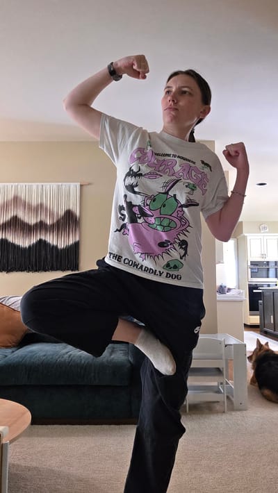 A woman with short dark hair is standing in a room and flexing her arms. She is wearing a light gray t-shirt with a colorful graphic of "The Cowardly Dog" cartoon character and the text "WELCOME TO NOWHERE". She is also wearing black pants and white socks. The woman is flexing her biceps with her fists clenched and her face is looking towards the camera with a playful expression.

The scene appears to be indoors, possibly in a living room or bedroom, based on the furniture visible in the background. A dark teal sofa is partially visible to the left, and a cream-colored wall with a piece of wall art hangs behind her. A wooden side table is also present. The floor is carpeted in a neutral tone. A small dog, possibly a German Shepherd mix, is lying on the carpet in the lower left corner of the frame. The room is well-lit, suggesting it could be daytime. There are no strong indicators of weather conditions.

The image is taken from a slightly low angle, and is rotated so that the subjects are sideways. The overall mood of the image is lighthearted and fun, with the woman's pose and expression conveying a sense of playful strength.