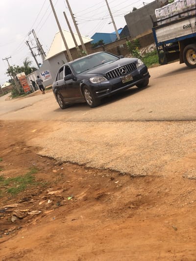 The image depicts a street scene in Hanwa, Nigeria. A black Mercedes-Benz SUV is parked on the side of the road, partially obstructing the lane. Adjacent to the car, a truck with a white bed is also parked. On the opposite side of the street, a building is visible with a sign that reads "Top Villa Suites Annex." A person is walking on the sidewalk in front of the building. Overhead, power lines crisscross the sky. The road is paved but shows signs of wear and tear. To the right of the pavement, there is unpaved terrain consisting of gravel and bare earth.
