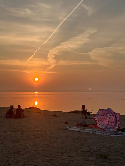 The image depicts a sunset scene on a sandy beach, likely in Surakovo, Russia, given the context. The sun, a bright orange orb, is low on the horizon, casting a warm, golden glow across the sky and water. A distinct contrail streaks across the sky, indicating aircraft activity. In the foreground, two individuals are seated on the sand, facing the water, appearing to be engaged in conversation or enjoying the view. Nearby, beach paraphernalia, including a colorful umbrella, blankets, and chairs, are set up, suggesting a leisurely outing. The water stretches out to the horizon, reflecting the sunset's colors and appearing calm. A series of small buoys or markers can be seen as a dotted line across the water. The sandy terrain of the beach dominates the foreground, with subtle textures and shadows.