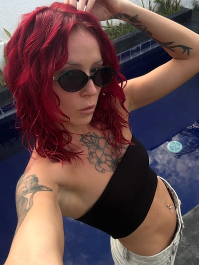 A young woman with red hair is lying down outdoors. She is wearing a black crop top, light-wash denim shorts, and round sunglasses. She has tattoos on her arms and chest, and a belly button piercing. She appears to be next to a blue pool or water feature. The location is outdoors in Dalung, Indonesia.