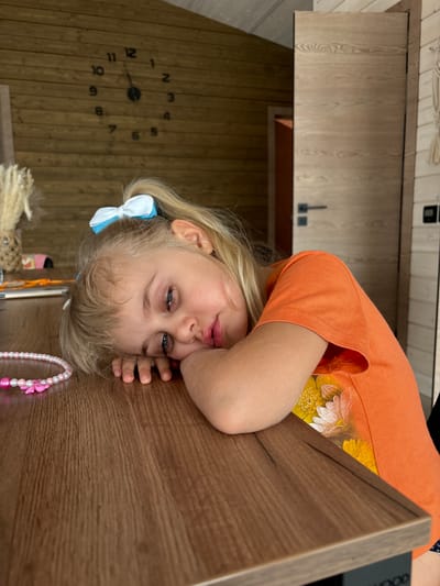 A young girl with blonde hair and blue eyes, wearing an orange t-shirt with a floral print, rests her head on a wooden table, looking directly at the camera with a pensive expression. A blue and white hair bow adorns her hair. Beside her on the table, a pink pearl necklace with flower charms is visible. In the background, a wooden wall features a large, minimalist clock with black numbers and hands, displaying the time as approximately 10:08. The setting appears to be indoors, with wooden paneling on the walls and a wooden cabinet or counter visible. The overall lighting suggests daytime.