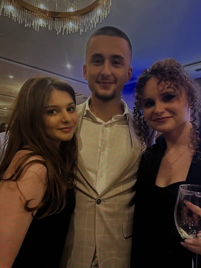 The image shows three young adults posing for a photograph. A man is positioned in the center, with a woman on his left and another woman on his right. The man is wearing a light-colored suit with a checkered pattern, a white collared shirt, and has short, dark hair. The woman on his left has long, brown hair and is wearing a black top. The woman on his right has curly, light brown hair, is wearing a black top, and is holding a wine glass. The background appears to be an indoor event space, possibly a restaurant or banquet hall, with a chandelier visible overhead. The lighting is dim and suggests an evening setting. The location is not specified as Novi Sad, Serbia.