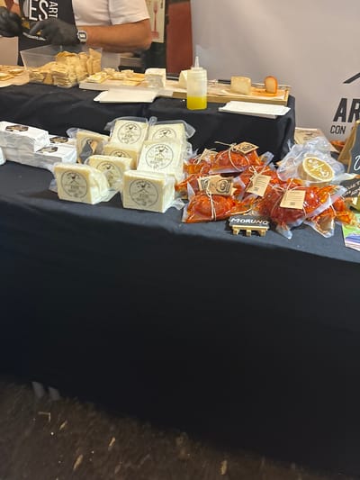 The image shows a display of artisanal cheeses and cured meats on a black tablecloth-covered table at a food market. Several stacks of cubed cheeses, wrapped in clear plastic, are visible, each with a label bearing the name "Jerusalem Exide." Additionally, various packages of chorizo or similar cured meats are displayed, labeled "MORUNO." Several cheese slices and a bottle of yellow sauce are on display for tasting. The table is located at Mercado Victoria in Córdoba, Spain. A person wearing a black glove, white shirt, and a watch can be seen on the left side of the table. A sign with text "Art CON" is partially visible in the background. The floor to the right of the table is unpolished concrete.