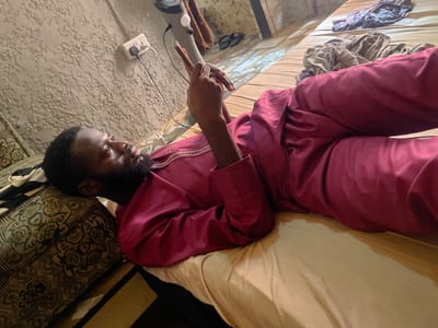A person is lying on their back on a bed, facing left. They are wearing a maroon-colored outfit. The person is holding a smartphone. There's a power outlet visible on the wall and a floor lamp. The location is likely indoors, possibly a residence in Lafia, Nigeria, but this is speculative. The bed has a tan sheet and an ornate headboard. Clothes are scattered on the bed in the background.