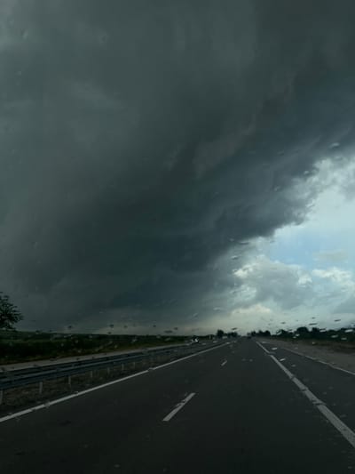 The view from a vehicle's windshield reveals a dark, stormy sky dominating the left side of the frame. Raindrops are visible on the glass. To the right, a section of asphalt highway stretches into the distance with a painted white line marking lanes. A strip of greenery borders the road. The location is identified as Zlatina, Bulgaria. No people or specific interactions are visible.