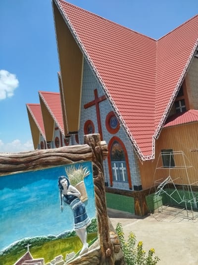 In K'Rèn, Vietnam, an angular, modern church building with a red tiled roof stands against a bright blue sky with a few scattered clouds. The building features a unique architectural design with sharply peaked roof sections and decorative metal trim along the edges. The facade is a textured gray material with patterned gray tiles, accented by red crosses and circular stained-glass windows. The main entrance area features a large red cross on the facade. To the right, a section of the building is clad in warm-toned wood. In the foreground, a decorative relief sculpture is visible, depicting a woman in traditional clothing carrying a basket of grain on her back, with a stylized landscape and a small church in the background. Yellow wildflowers grow at the base of the sculpture. The ground appears to be a paved courtyard.