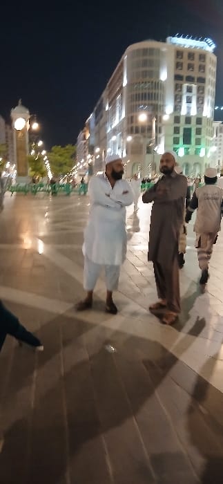 The image displays a nighttime scene within a well-lit, paved urban plaza in Medina, Saudi Arabia, identifiable by the distinctive architecture and the "ZAM ZAM" uniform, consistent with the area surrounding Al-Masjid an-Nabawi (The Prophet's Mosque).

In the mid-ground, two adult males stand conversing or observing, both with their arms crossed. The man on the left, of medium build with a dark beard, wears a white thobe, white shalwar, and a white taqiyah, with dark brown sandals. The man on the right, also of medium build with a dark beard, wears a dark brown thobe and matching shalwar, with dark footwear. In the right background, a third male figure is seen walking away, clad in a light-colored uniform (khaki/beige) with "ZAM ZAM" printed on the back. A partial view of a dark-clad leg and foot belonging to a fourth individual is visible in the extreme bottom left foreground.

The environment is illuminated by numerous artificial light sources, including streetlights and lights from buildings. A prominent light-colored clock tower with an illuminated clock face is visible in the far left background. To the right of the clock tower, a modern, multi-story building with illuminated windows stands in the background. Green barriers or bollards line segments of the paved plaza. The ground consists of large, light-colored rectangular paving tiles.