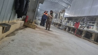 Two individuals stand in a large industrial space with concrete ceilings and metal structures. One person wears camouflage clothing, while the other is in blue attire with an orange vest. Visible elements include shelving with objects, metal beams, and corrugated metal siding. The scene is well-lit but the image has been rotated.