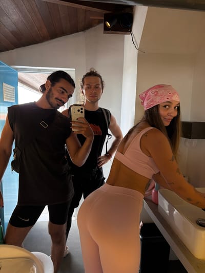 In a dimly lit room with a wooden ceiling, three individuals are reflected in a large mirror. On the left, a man with dark, pulled-back hair and a beard, wearing a sleeveless black t-shirt and black shorts, holds a smartphone to take a selfie. To his right, another man with medium-length, light brown hair, wearing a black t-shirt, stands looking towards the mirror. In the foreground, a woman with long brown hair tied up with a pink bandana and wearing a pink sports bra and pink leggings, leans towards a sink. Her back is to the camera, and she appears to be interacting with the sink. The environment appears to be a bathroom, with a sink and a black trash can visible. The reflection of a blue door with a vent can be seen on the left side of the mirror.