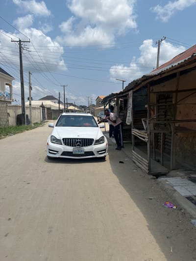 The image captures a street scene in Iranla, Nigeria, under a bright blue sky with scattered white cumulus clouds. A white Mercedes-Benz C-Class sedan, displaying a Lagos State license plate "AAA-860KA," is stationary on an unpaved dirt road, facing the viewer. A red object rests on its dashboard. To the immediate right of the vehicle, a male individual wearing a light pink short-sleeved polo shirt and dark track pants leans towards the passenger side of the car, with his right hand near the window frame and his left hand near his face, possibly gesturing or holding an object. A second male, partially obscured behind the first, stands further back near the car's rear passenger door. The right side of the road is bordered by a series of makeshift commercial structures or stalls constructed from wooden frames and topped with corrugated metal roofs, some exhibiting signs of rust and disrepair. Litter, including plastic wrappers, is visible on the dirt ground near these structures. The left side of the road features more developed residential buildings with concrete walls, one showing a light blue balcony railing, and perimeter walls topped with security elements. Multiple wooden utility poles with numerous overhead power lines line both sides of the street, extending into the background. A solar-powered street light is affixed to one of the utility poles on the right.