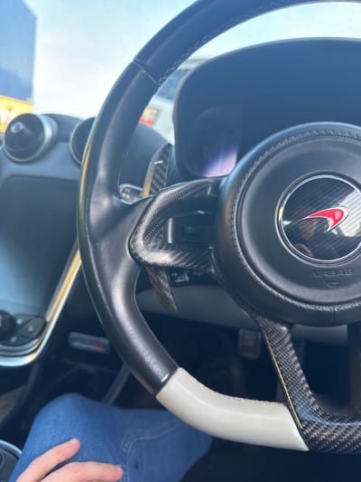 The image shows the interior of a vehicle, specifically the steering wheel. The steering wheel has a black leather grip, carbon fiber accents, and a white grip section. The central emblem is a red and silver logo within a black circle marked "AIRBAG." Paddle shifters are visible behind the wheel, constructed of carbon fiber. The dashboard is partially visible, including a control panel with buttons. A person's leg in blue jeans and hand are visible to the right. The city location Great Yarmouth and United Kingdom do not feature in this picture.